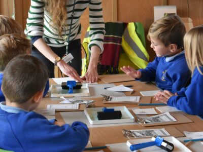 A group of KS1 children sitting at a table watching a printmaking demonstration.